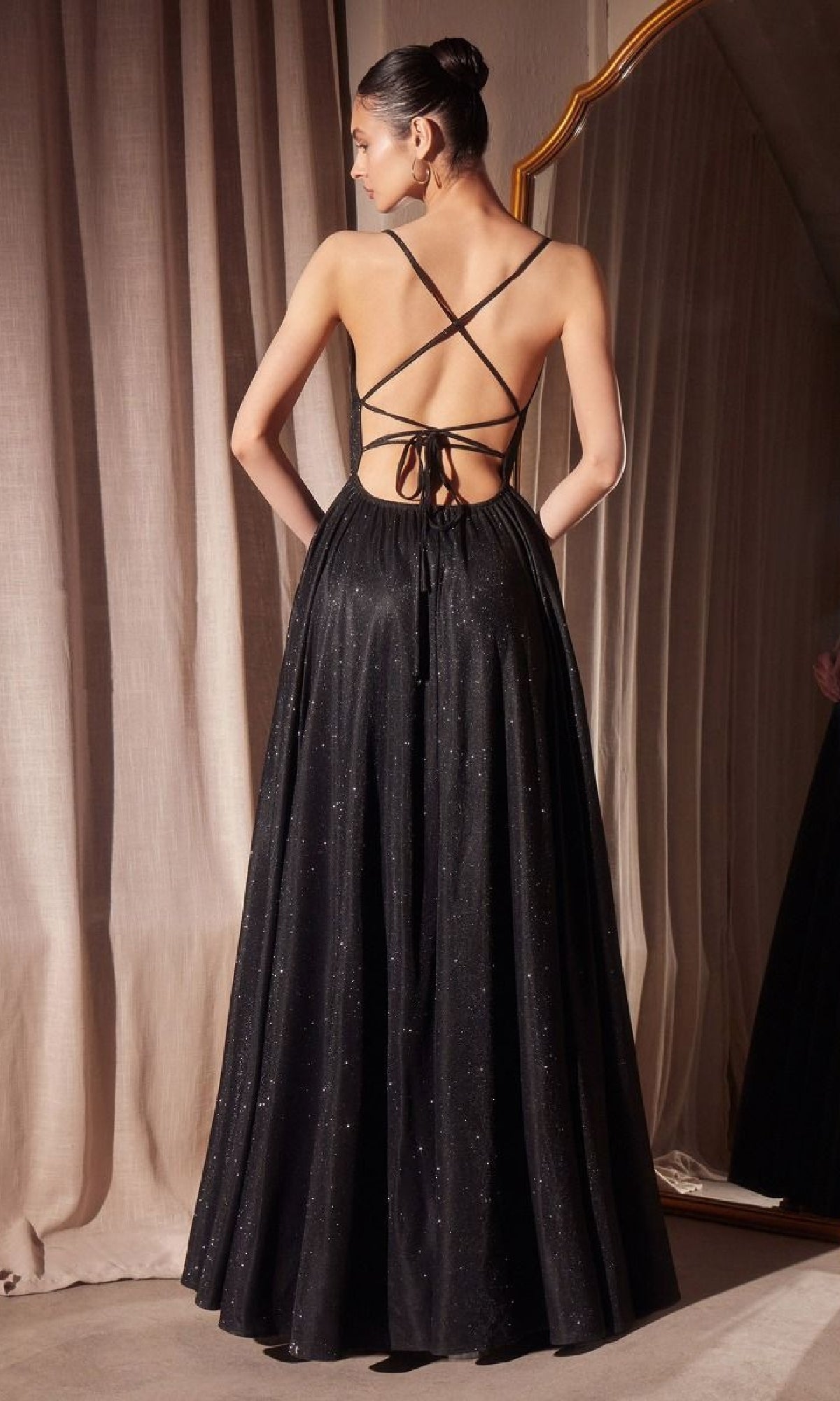 A woman faces away from the camera in the Strappy-Back Glitter Prom Gown: Amoris T214, featuring crisscross straps and a full skirt. A mirror and beige curtains are visible in the background.